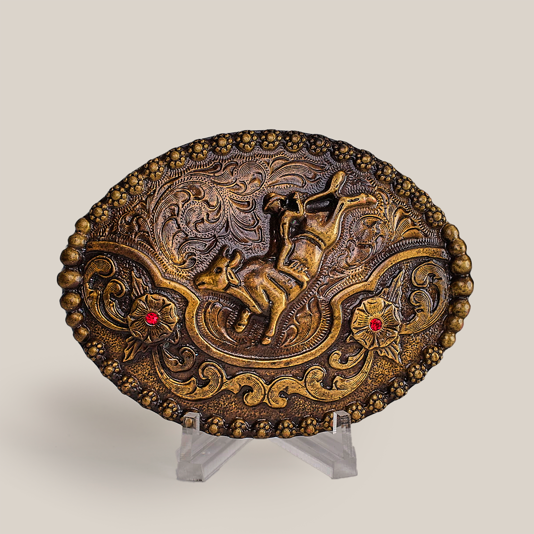 The Ruby Bull Rider Buckle is an ornate, oval bronze piece with an embossed horse amid intricate floral and swirl designs, echoing Texas-style buckles. Displayed on a clear stand against a simple backdrop, it exudes rustic charm and craftsmanship.
