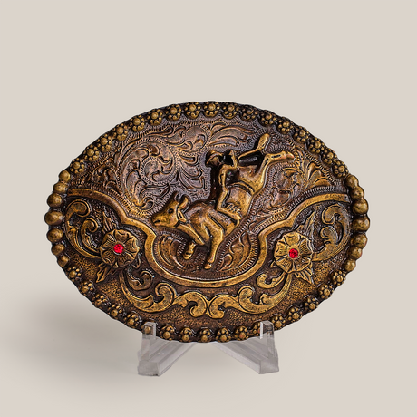 The Ruby Bull Rider Buckle is an ornate, oval bronze piece with an embossed horse amid intricate floral and swirl designs, echoing Texas-style buckles. Displayed on a clear stand against a simple backdrop, it exudes rustic charm and craftsmanship.
