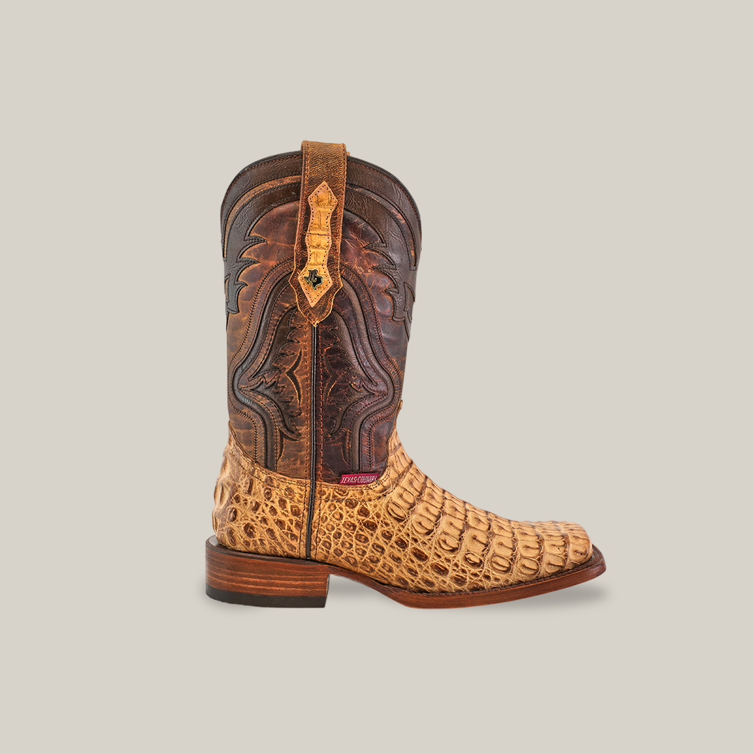 The Exotic Caiman Hornback - Antique - Rodeo Toe cowboy boot showcases a textured design with a brown upper featuring intricate stitching and an authentic caiman leather lower in a tan crocodile skin pattern. Its wooden sole and heel create a striking look on a plain white background.