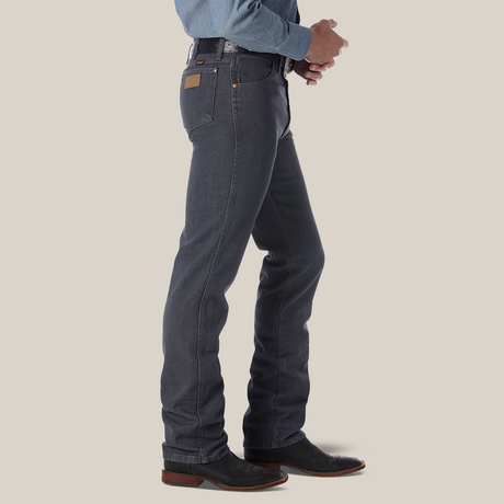 A person stands against a plain background wearing premium Wrangler Cowboy Cut Slim Fit Jean - 0936CHG in dark denim, paired with a blue shirt and black cowboy boots, captured from a shoulder-down side profile to highlight the fit and style.