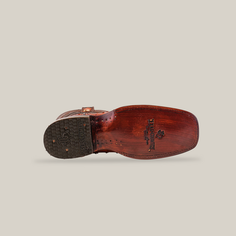 The image features the sole of a single Exotic Ostrich - Brandy - Rodeo Toe cowboy boot, artistically positioned on its side. Crafted from smooth brown ostrich leather, it showcases exceptional craftsmanship, visible logos, and a textured heel for grip against the plain light background.