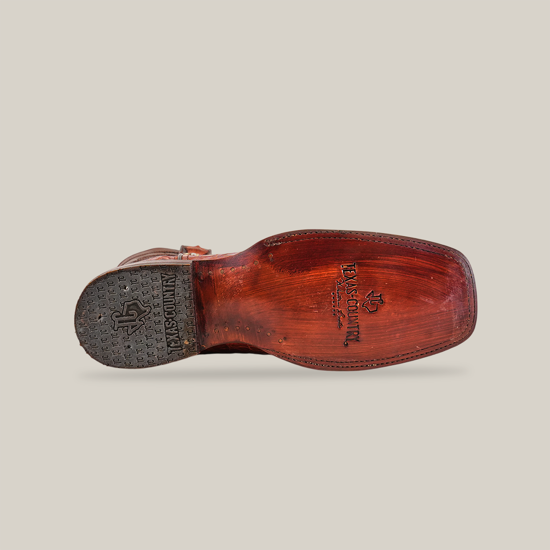 The image shows the underside of a well-worn Exotic Caiman Hornback - Brandy - Rodeo Toe boot. The textured, luxurious leather sole displays branding and logos. The heel features more branded accents, and the scuffed, genuine caiman leather highlights its authentic craftsmanship.