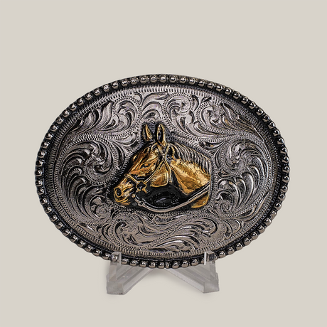 The Stud Horse Buckle is an ornate, oval-shaped Texas-style creation in alpaca silver, featuring intricate western scrollwork with a central gold horse head design against a neutral background.