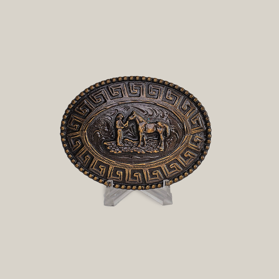 The Horsemen Buckle is an oval alpaca silver plate with an intricate Greek key border, showcasing a central relief of a figure leading a horse amidst nature elements, designed in western style. It is elegantly displayed on a clear stand against a gray background.