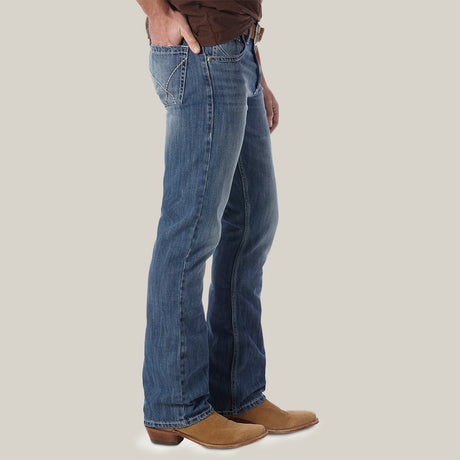 A person, shown from shoulders down, wears a brown shirt, premium denim blue jeans with decorative back pocket stitching, and Mens Wrangler 20X No. 42 Vintage Bootcut Jeans. They stand against a plain light gray background.