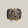 The Charro Horse Buckle features a Texas-style rectangular design with a horse head motif in rich gold tones, accentuated by swirling patterns and a Greek key border. Displayed on a clear stand against a gray backdrop, it embodies classic western designs.