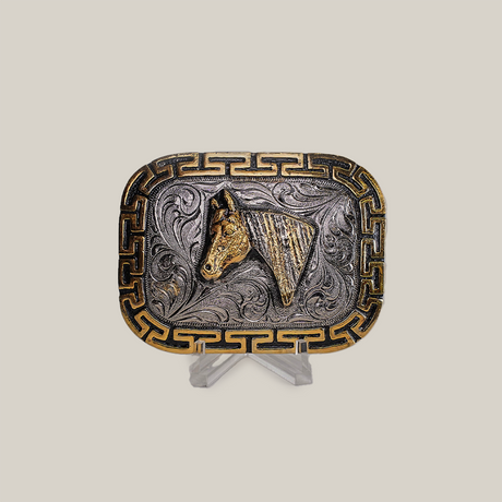 The Charro Horse Buckle features a Texas-style rectangular design with a horse head motif in rich gold tones, accentuated by swirling patterns and a Greek key border. Displayed on a clear stand against a gray backdrop, it embodies classic western designs.