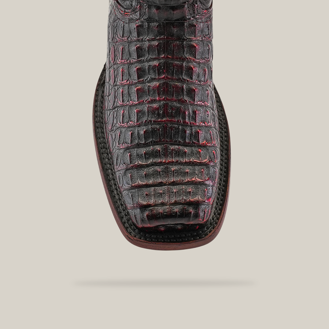A close-up of the Exotic Caiman Hornback - Black Cherry boot showcases its black and red crocodile pattern with a square toe, crafted from genuine caiman leather. White stitching around the sole highlights expert craftsmanship against a pristine white background.