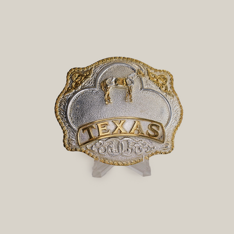 The Texas Horse Buckle boasts a Texas theme with western designs in gold and alpaca silver, featuring a star, horse, and TEXAS embossed in gold. Its elegantly displayed on a clear stand against a plain background.