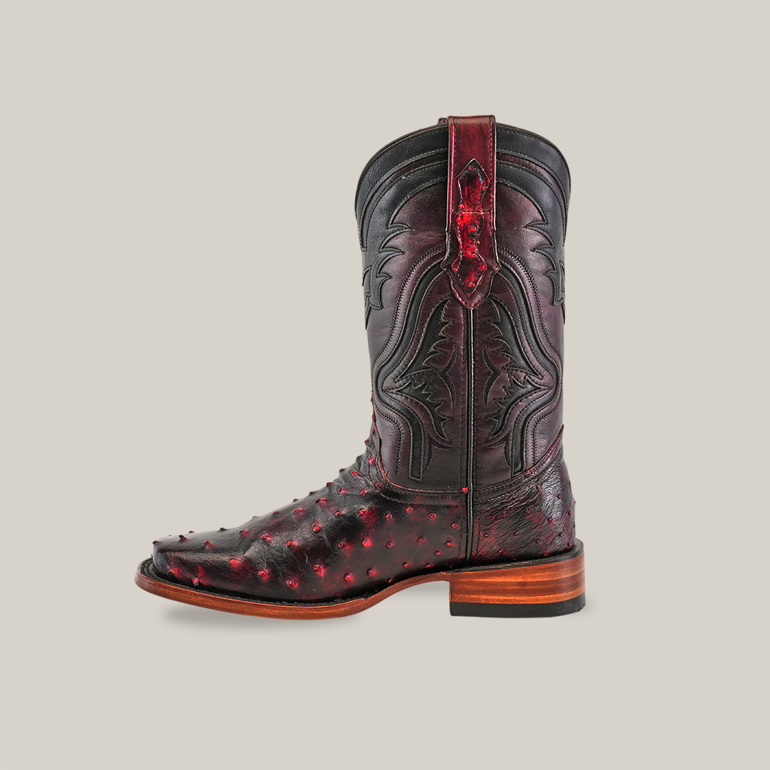 The Exotic Ostrich - Black Cherry cowboy boot, crafted from genuine ostrich leather, features a wooden heel and intricate red-accented patterns. Its textured surface and ornamental detailing with a stylish rodeo toe stand out against a plain white background.