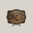 The Ruby Rooster Buckle is an antique brass belt buckle featuring intricate engravings in a classic western design. It showcases a raised rooster centerpiece encircled by floral patterns and embellished with two red gemstone accents, reminiscent of Texas-style buckles. Displayed elegantly on a clear stand.