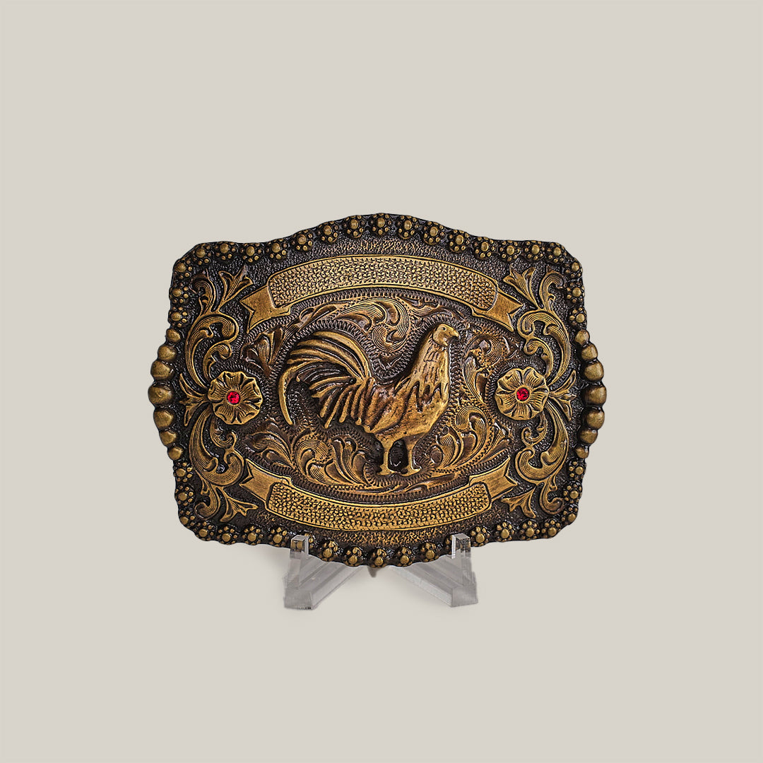 The Ruby Rooster Buckle is an antique brass belt buckle featuring intricate engravings in a classic western design. It showcases a raised rooster centerpiece encircled by floral patterns and embellished with two red gemstone accents, reminiscent of Texas-style buckles. Displayed elegantly on a clear stand.