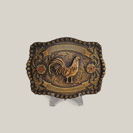 The Ruby Rooster Buckle is an antique brass belt buckle featuring intricate engravings in a classic western design. It showcases a raised rooster centerpiece encircled by floral patterns and embellished with two red gemstone accents, reminiscent of Texas-style buckles. Displayed elegantly on a clear stand.