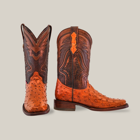 The Exotic Ostrich - Brandy - Rodeo Toe boots, crafted from textured brown ostrich leather with intricate embroidery, are displayed against a white background. One boot stands upright while the other tilts to showcase its sole, highlighting the exceptional craftsmanship.