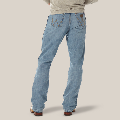 A rear view shows a person wearing Mens Wrangler Retro Relaxed Fit Bootcut Jeans - WRT20CR, in light blue, paired with a gray long-sleeve shirt. The jeans display back pockets with decorative stitching and a leather patch on the waistband, set against a light background.