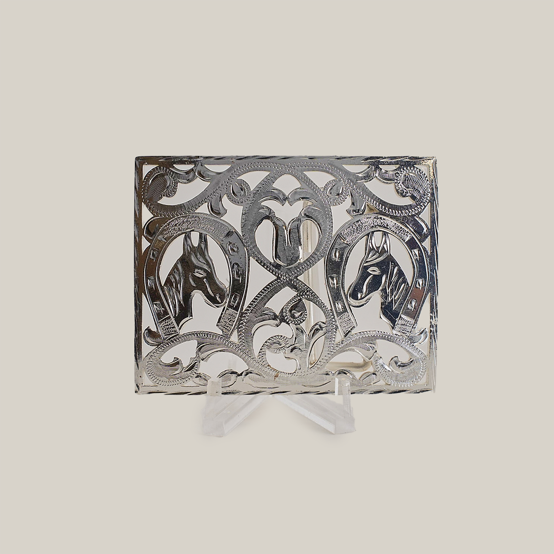 The Double Horseshoe Horse Buckle is an ornate alpaca silver plaque with cut-out patterns, showcasing two horse head profiles in circular designs. It features intricate swirling motifs and a central heart shape, displayed on a transparent stand against a plain background, echoing western designs.