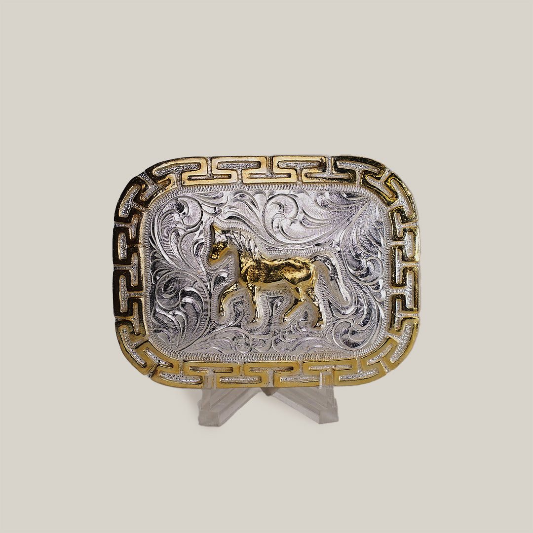 The Charro Full Horse Buckle showcases a rectangular alpaca silver design with intricate western engravings, a central gold horse motif, and golden geometric borders. Elegantly presented on a clear stand, it embodies classic Texas-style craftsmanship.