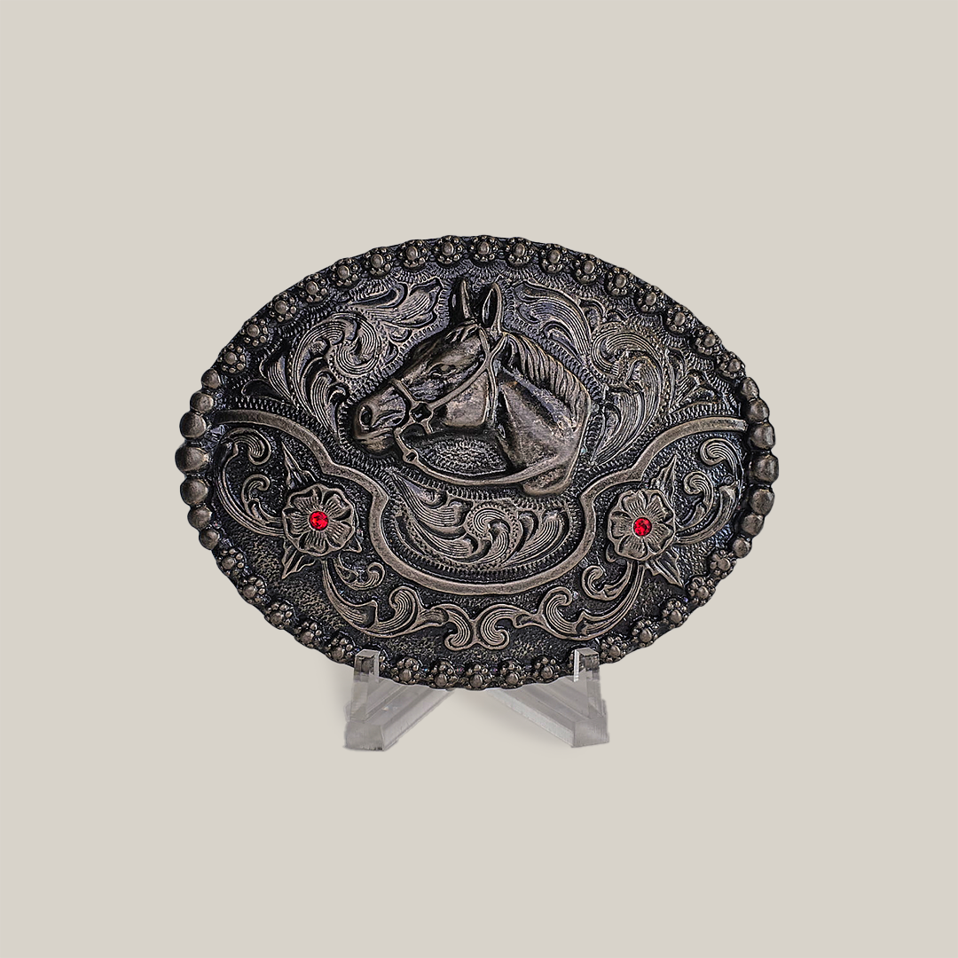 The Ruby Horse Buckle is an alpaca silver oval belt buckle with a raised horse head motif, two red gems, and Texas-style swirls. Its displayed on a clear stand against a neutral background.