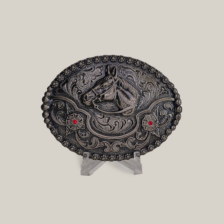 The Ruby Horse Buckle is an alpaca silver oval belt buckle with a raised horse head motif, two red gems, and Texas-style swirls. Its displayed on a clear stand against a neutral background.