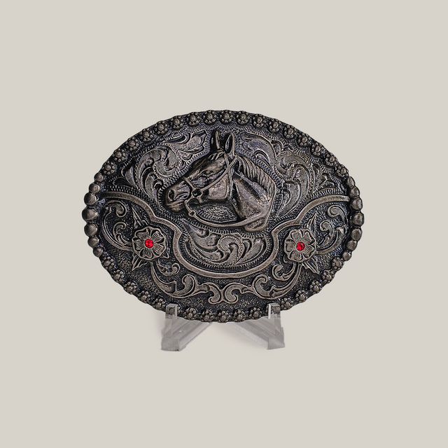 The Ruby Horse Buckle is an alpaca silver oval belt buckle with a raised horse head motif, two red gems, and Texas-style swirls. Its displayed on a clear stand against a neutral background.
