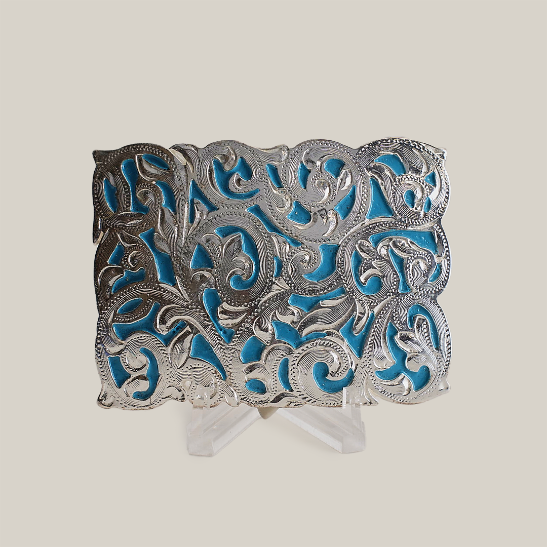 The Charro Turquoise Buckle is an ornate alpaca silver piece featuring intricate scrolling patterns on a vibrant blue background. Its cut-out design showcases exceptional craftsmanship with highlighted curves and flourishes, reminiscent of classic Texas-style buckles, displayed upright on a stand.