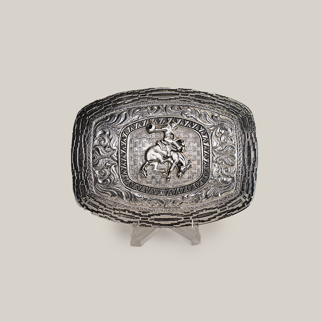 The Charro Floral Bull Rider Buckle, a rectangular Texas-style belt buckle made from alpaca silver, showcases an intricate design with a bull rider at its center, surrounded by ornate floral and scroll patterns. Displayed on a clear stand against a plain background, it exudes Western charm.