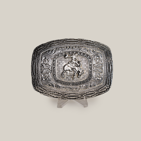 The Charro Floral Bull Rider Buckle, a rectangular Texas-style belt buckle made from alpaca silver, showcases an intricate design with a bull rider at its center, surrounded by ornate floral and scroll patterns. Displayed on a clear stand against a plain background, it exudes Western charm.