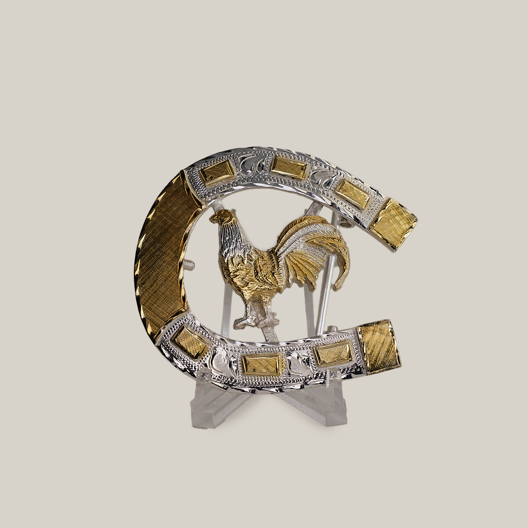 The HorseShoe Rooster is a decorative silver and gold horseshoe with ornate detailing, showcasing a golden rooster centrally. It features intricate patterns and textures, reminiscent of western designs, and is displayed on a transparent stand against a plain background.