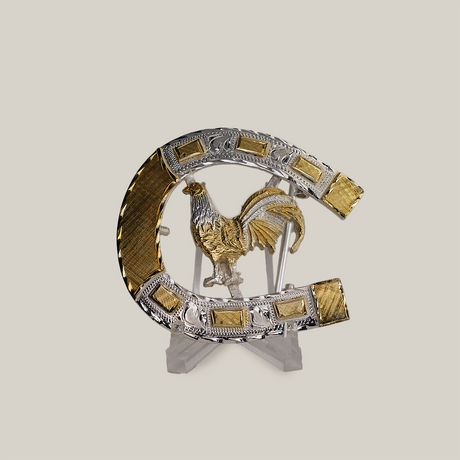 The HorseShoe Rooster is a decorative silver and gold horseshoe with ornate detailing, showcasing a golden rooster centrally. It features intricate patterns and textures, reminiscent of western designs, and is displayed on a transparent stand against a plain background.