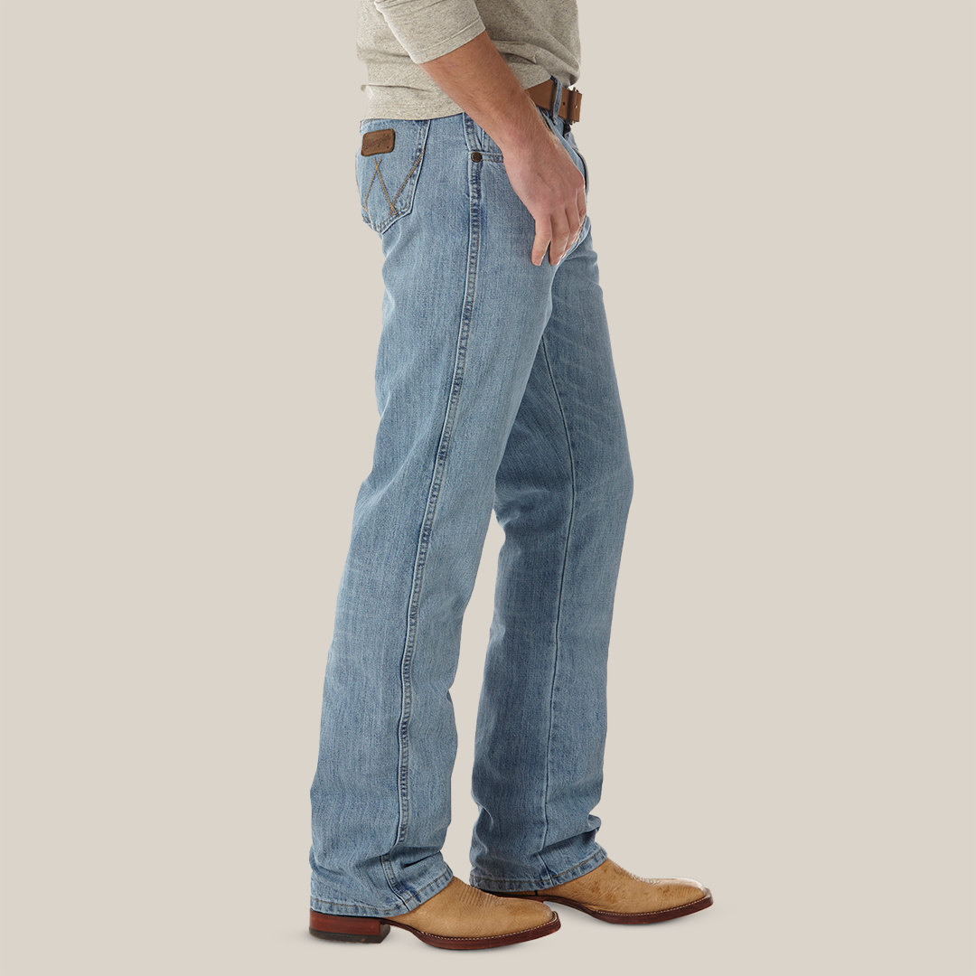 A person in light blue Mens Wrangler Retro Relaxed Fit Bootcut Jeans (WRT20CR) with a brown leather patch and subtle back pocket embroidery, paired with a brown belt, beige cowboy boots, and a light gray long-sleeve shirt, stands against a plain background.
