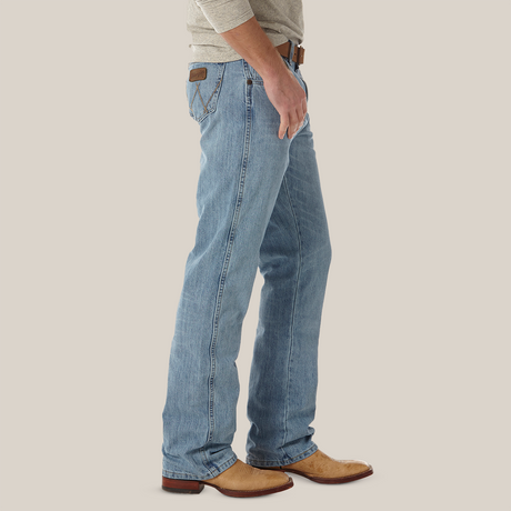 A person in light blue Mens Wrangler Retro Relaxed Fit Bootcut Jeans (WRT20CR) with a brown leather patch and subtle back pocket embroidery, paired with a brown belt, beige cowboy boots, and a light gray long-sleeve shirt, stands against a plain background.