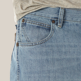 A close-up shows a person wearing the Mens Wrangler Retro Relaxed Fit Bootcut Jean - WRT20CR with a gray shirt. The light blue denim features bootcut style, front pocket with copper rivets, visible stitching, and a classic, slightly worn texture.