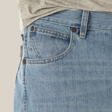 A close-up shows a person wearing the Mens Wrangler Retro Relaxed Fit Bootcut Jean - WRT20CR with a gray shirt. The light blue denim features bootcut style, front pocket with copper rivets, visible stitching, and a classic, slightly worn texture.