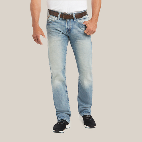 Someone in a white shirt, light blue M7 Rocker Stretch Stirling Stackable Straight Leg Jean - Shasta (10031997), and black shoes stands against a plain backdrop. The slightly faded jeans with stretch fabric are complemented by the brown belt held in one hand. The image is cropped at the shoulders.