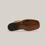 Bottom view of a worn Exotic Caiman Hornback - Orix boot with a square toe, crafted from premium leather. The sole shows visible wear with engraved text and a logo featuring a small figure and decorative patterns on the heel, set against a white background to showcase its rustic charm.