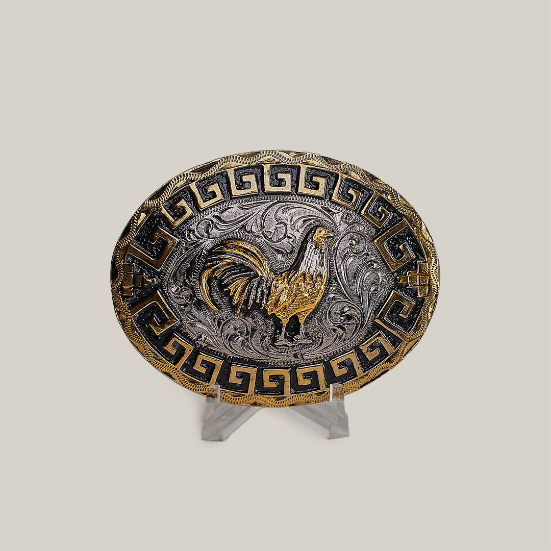 The Charro Rooster Buckle is an oval belt buckle with a western design, featuring a gold rooster on a silver background with swirling patterns. It has a Greek key motif in gold and black on the border. Made from alpaca silver, it is displayed on a clear stand against a gray background.