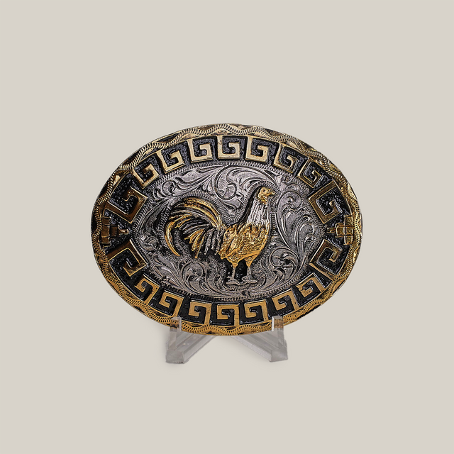 The Charro Rooster Buckle is an oval belt buckle with a western design, featuring a gold rooster on a silver background with swirling patterns. It has a Greek key motif in gold and black on the border. Made from alpaca silver, it is displayed on a clear stand against a gray background.