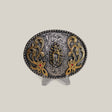 The Virgen Maria Buckle is an ornate oval-shaped belt buckle with intricate western designs, detailed engravings, a central figure, and floral patterns. It features gold accents and red stones, displayed on a clear stand against a neutral background.