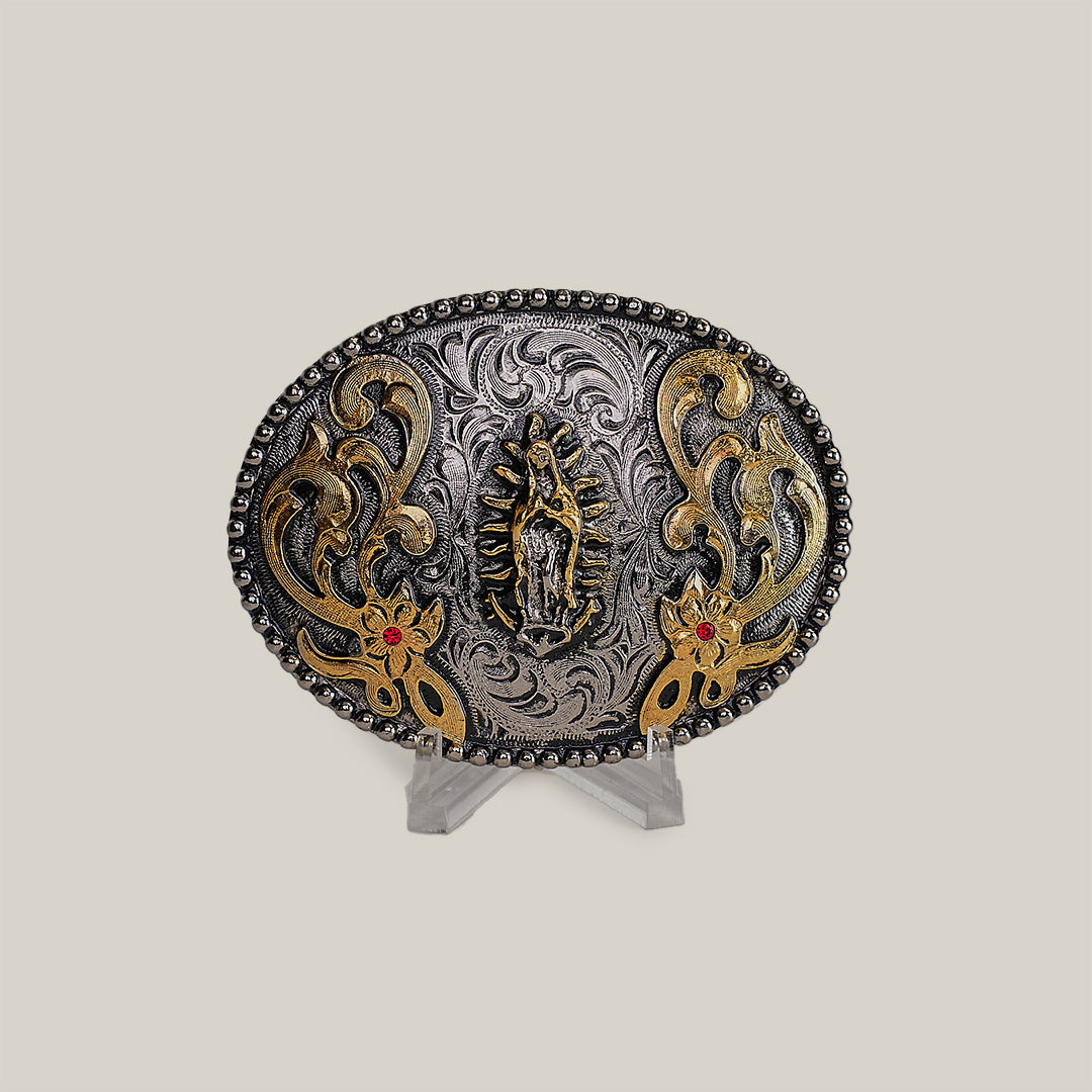 The Virgen Maria Buckle is an ornate oval-shaped belt buckle with intricate western designs, detailed engravings, a central figure, and floral patterns. It features gold accents and red stones, displayed on a clear stand against a neutral background.