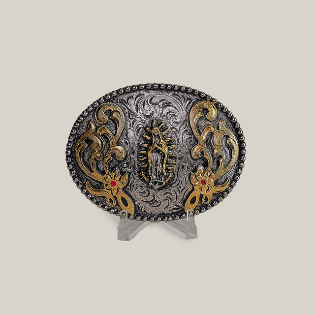 The Virgen Maria Buckle is an ornate oval-shaped belt buckle with intricate western designs, detailed engravings, a central figure, and floral patterns. It features gold accents and red stones, displayed on a clear stand against a neutral background.