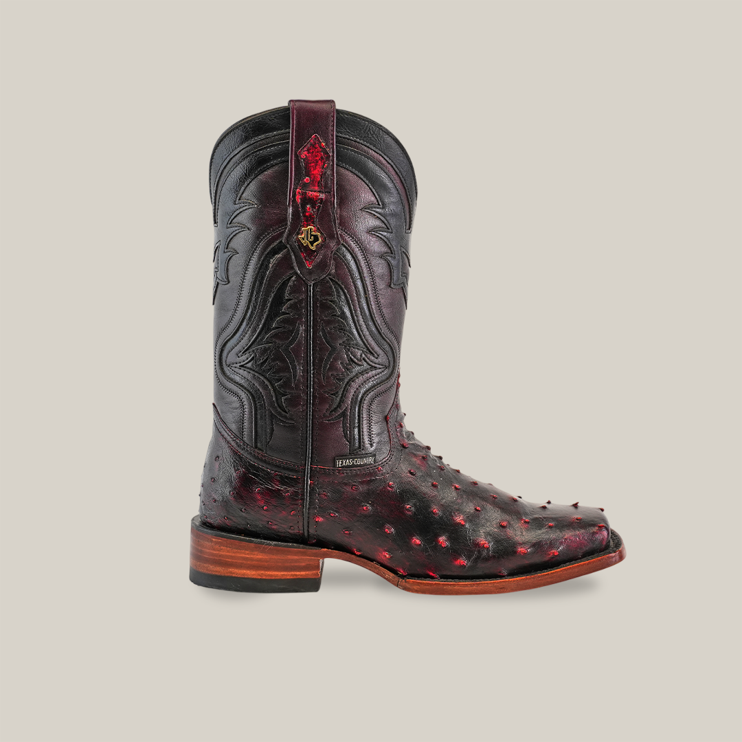 The Exotic Ostrich - Black Cherry boot is crafted from genuine ostrich leather, featuring red and black patterns, a dark leather shaft, a wooden heel, ornate stitching with red accents on the lower part, and a classic rodeo toe.