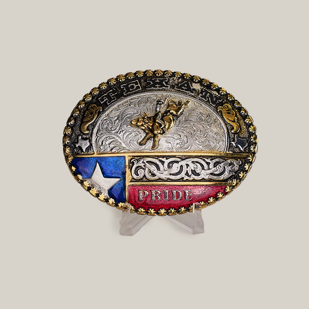 The Texas Bull Rider Buckle is an oval belt buckle with a striking blend of gold and alpaca silver, displaying TEXAN at the top. It features a cowboy on horseback with PRIDE, along with a Texas flag motif and star on a red and blue backdrop, elegantly showcased on a stand.
