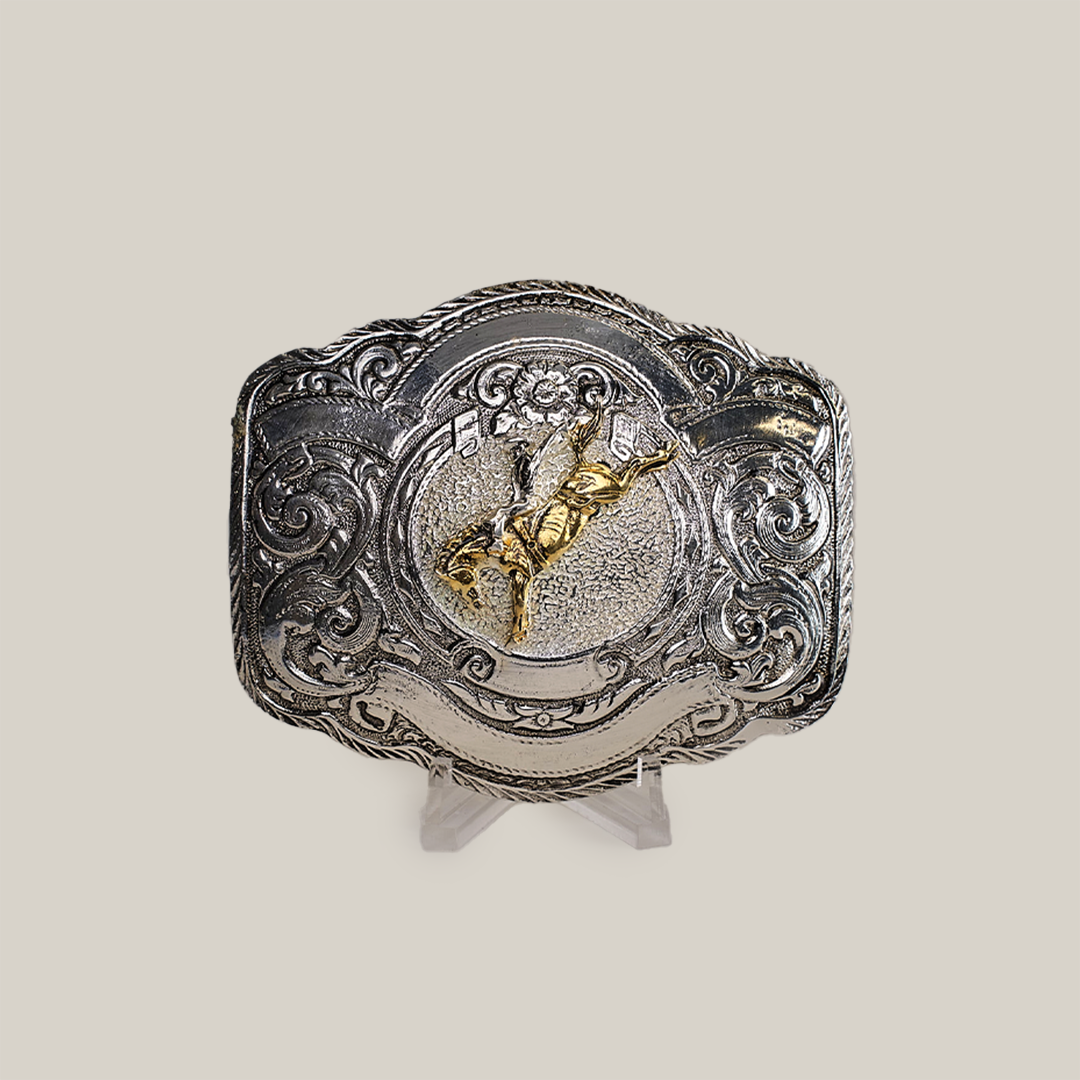 The Floral Bull Rider Buckle, crafted from alpaca silver, showcases intricate western designs and a raised gold-colored bull rider in the center. Its elegantly presented on a clear stand against a plain gray background.