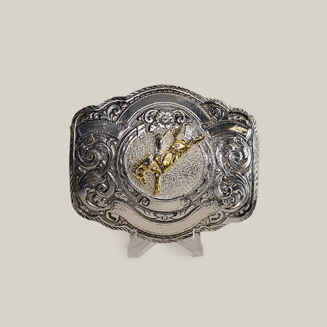 The Floral Bull Rider Buckle, crafted from alpaca silver, showcases intricate western designs and a raised gold-colored bull rider in the center. Its elegantly presented on a clear stand against a plain gray background.