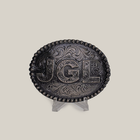 The JGL Buckle, an intricately designed oval piece made from alpaca silver, showcases bold JGL letters and ornate swirling patterns like Texas-style buckles. It is displayed on a clear stand against a plain backdrop.