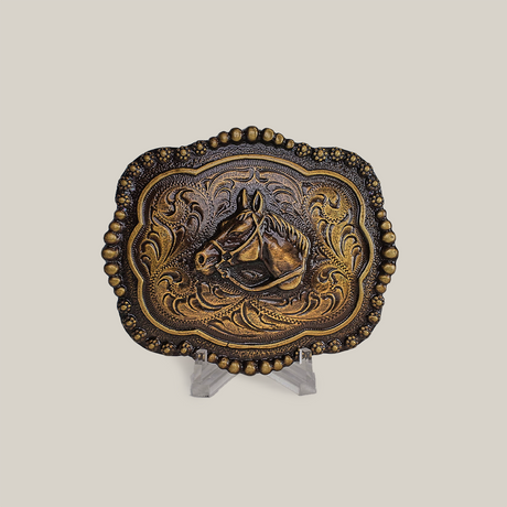 The Vintage Stud Horse Buckle in alpaca silver displays an embossed horse head and floral designs on a small stand. A beaded border enhances its shimmer in bronze and gold against a gray background.
