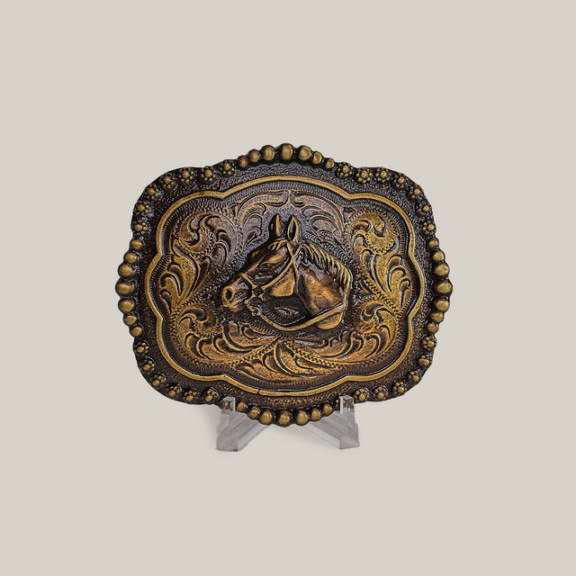 The Vintage Stud Horse Buckle in alpaca silver displays an embossed horse head and floral designs on a small stand. A beaded border enhances its shimmer in bronze and gold against a gray background.